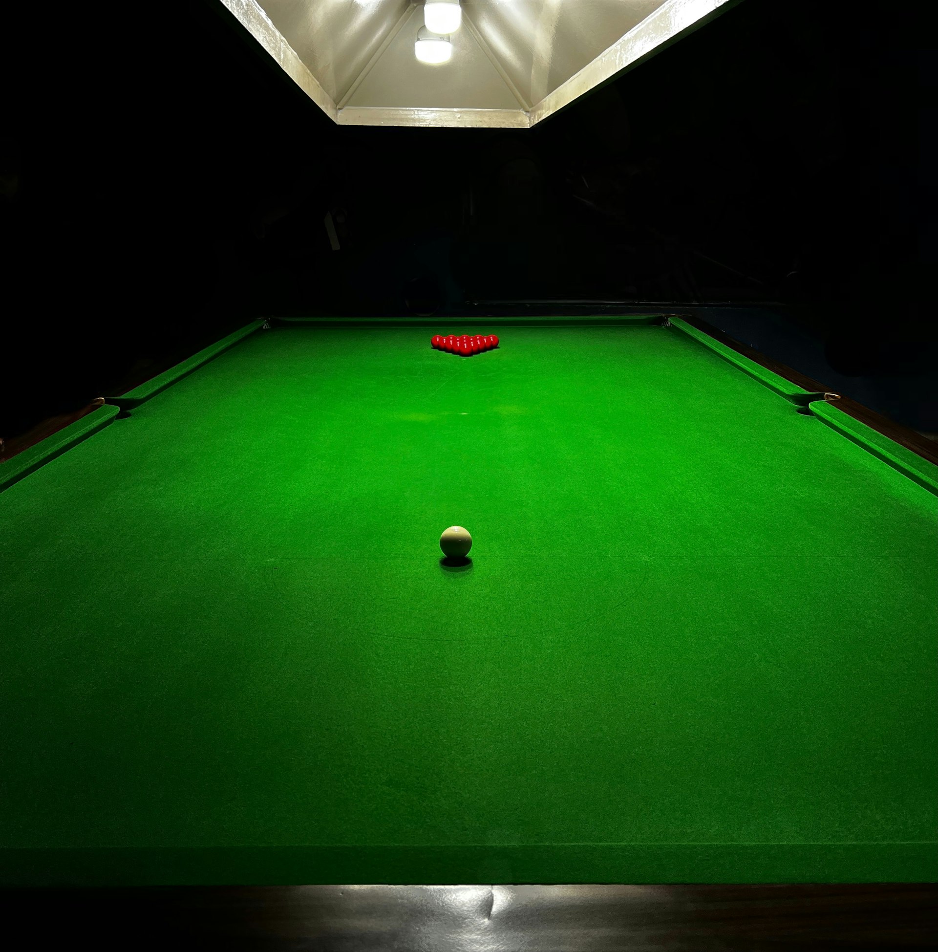 A snooker table lit from above in a dim club room, reds racked and the cue ball in play