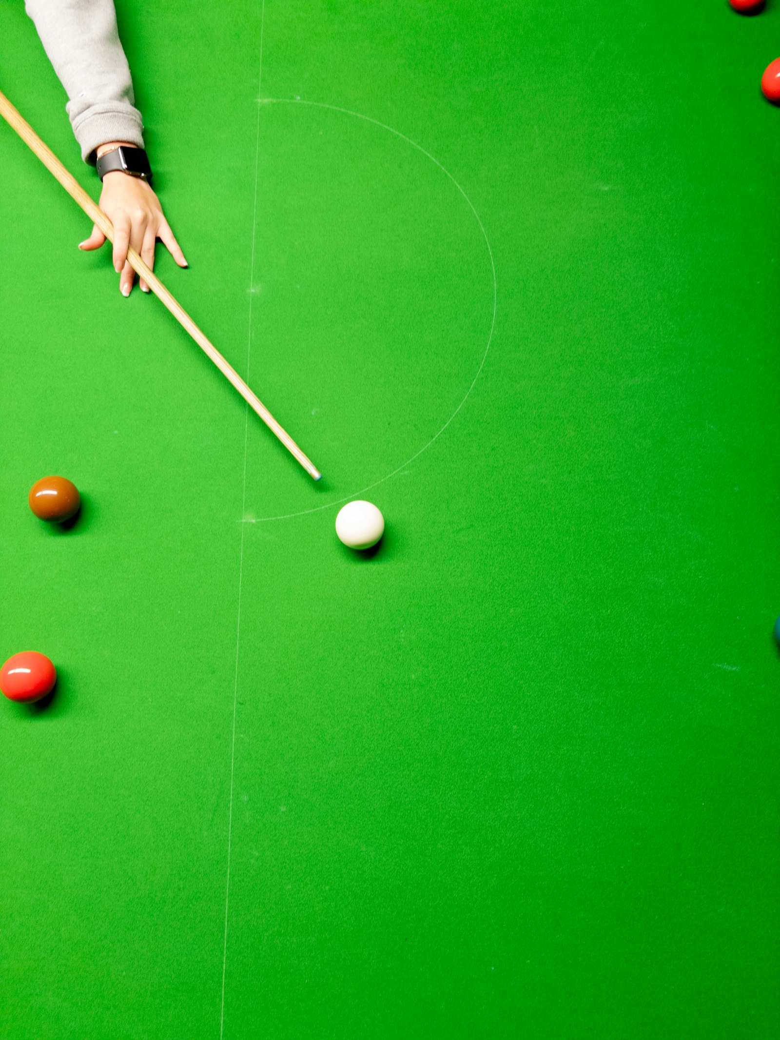 An aerial view of a snooker table with a player's hand addressing the cue ball near the baulk line, reds and a brown ball nearby